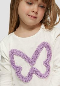 Weißes Hemd mit langen Ärmeln, das ein großes, strukturiertes lila Schmetterlingsdesign zeigt, das aus gerafftem Stoff entlang des Umrisses besteht.