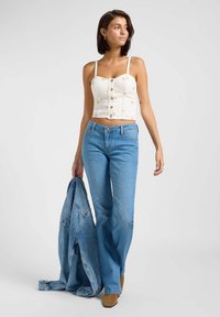 Hvit kort topp med knappelukking og tynne stropper, kombinert med lys blå flagrende jeans. En denimjakke er lagt over den ene armen.