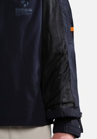 Chaqueta navy con mangas de malla negra en contraste. Cuenta con un logotipo gráfico en el pecho, cierre de cremallera y puños ajustables.