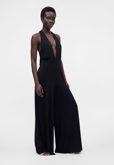 Femme portant une combinaison noire sans manches avec un profond col en V et un pantalon large, debout devant un fond clair uni.