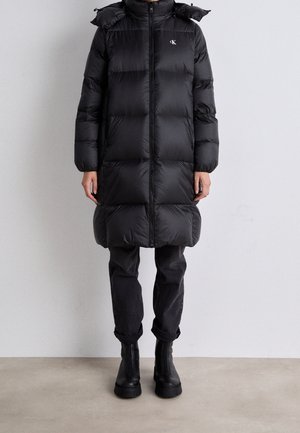 Personne portant un long manteau doudoune noir avec fermeture éclair, poches latérales, pantalon noir retroussé et bottines noires sur un fond uni.