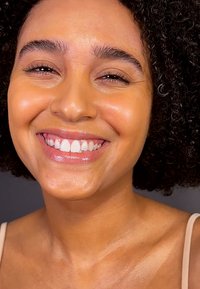 Leende person med lockigt mörkt hår, slät hy och naturlig makeup; bär en ljus topp, närbild som framhäver ansiktsdragen.