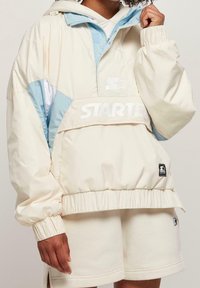Anorak ligero en crema y azul con dobladillo ajustable, puños elásticos y detalles del logo en el pecho y la manga. Tejido suave y liso.