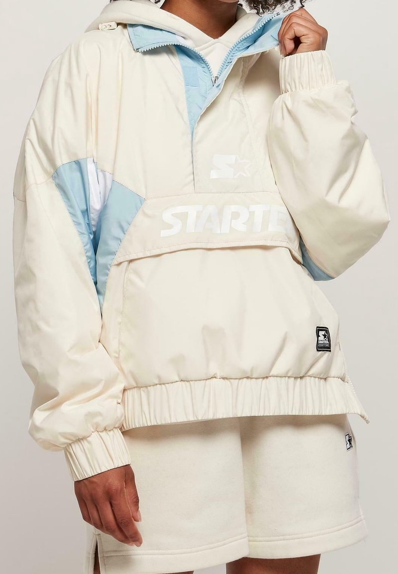 Anorak ligero en crema y azul con dobladillo ajustable, puños elásticos y detalles del logo en el pecho y la manga. Tejido suave y liso.