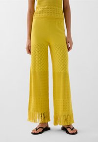 Pantalons larges en tricot jaune avec des motifs texturés, des détails en dentelle et des franges au niveau de l'ourlet. Portés avec des tongs noires.