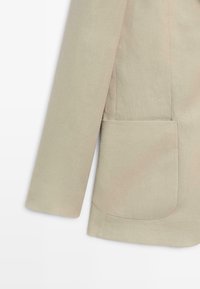 Mouw en onderste helft van een beige blazer met een groot afgerond voorvak met zichtbare stiksels op een effen achtergrond.
