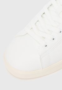 Adidași albi cu un superior sintetic neted și texturat, bot rotund și șireturi albe plate. Talpă din cauciuc cu accente bej deschis pe margine.