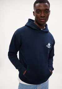 Hanorac bleu marin cu buzunar frontal și logo, fabricat din material moale. Prezintă o croială lejeră și glugă cu șnur. Purtați peste un strat alb.