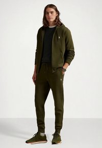 Zeleni bombažni pulover s kapuco in usklajene hlače z logotipom, oblečeni preko temne majice. Superge v ujemajoči se barvi dopolnjujejo obleko.