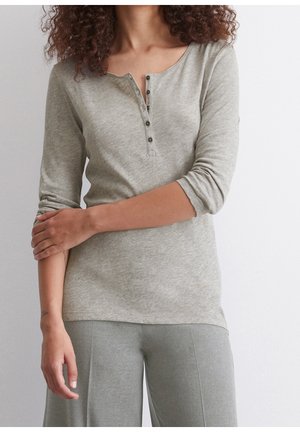 Femme aux cheveux bouclés portant un haut henley à manches longues gris clair avec des boutons et un pantalon ample assorti, se tenant devant un mur blanc.