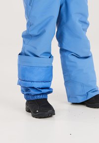 Blå isolerade snöbyxor med samlade benslut och kontrasterande röd sömnad, kombinerat med svarta vattentäta stövlar. Hållbart tyg för vinteranvändning.