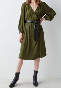 Femme portant une robe midi à manches longues olive avec une large ceinture noire nouée et des bottines noires, debout devant un fond uni.