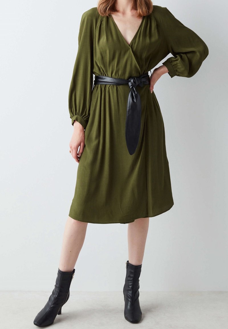 Femme portant une robe midi à manches longues olive avec une large ceinture noire nouée et des bottines noires, debout devant un fond uni.