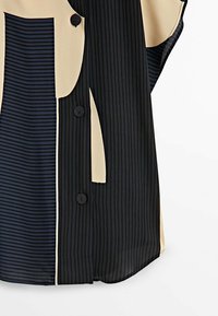 Blouse rayée noire et crème avec fermeture à boutons ; présente des largeurs de rayures variées et une texture lisse. Sections de couleurs contrastantes notables.