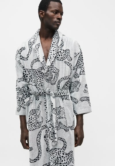 Άνδρας που φοράει ανοιχτό γκρι ρόμπα με μαύρο λεοπάρ μοτίβο, δεμένη στη μέση, κοιτάζοντας πλάγια σε μονόχρωμο φόντο.