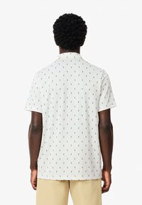 Valkoinen lyhythihainen poolopaita, jossa on toistuva laivastonsininen kuvio pienistä hahmoista. Sisältää normaalin kauluksen ja pehmeän kankaan tekstuurin.