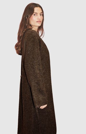 Vestido largo marrón de punto con acabado texturizado, con hombros caídos y un ajuste suelto, llevado por una modelo de perfil.
