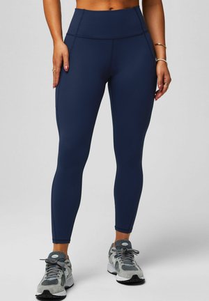 Leggings taille haute bleu marine en tissu lisse et extensible avec des coutures contrastantes. Associés à des chaussures de sport grises avec des détails en mesh.