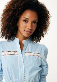 Ljusblå randig blus med hög krage, spetsdetaljer och knäppningar. Puffärmar med delikata spetsaccenter på bröstet.