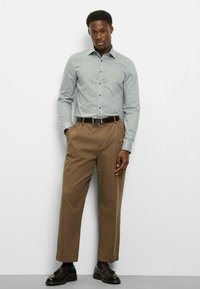 Hellblau gemustertes Hemd mit Knöpfen, kombiniert mit braunen, plissierten Hosen. Das Hemd hat einen Kragen, und die Hose ist locker geschnitten. Schwarze Schuhe vervollständigen den Look.