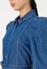 Vestido de ganga com gola, mangas bufantes e botões frontais. O tecido é azul claro com uma textura suave e detalhes mínimos.