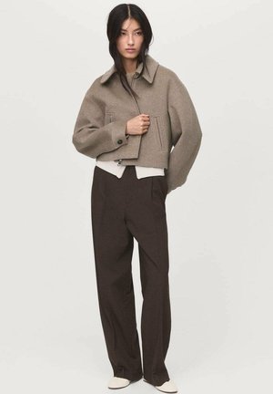 Veste en laine beige courte avec un large col et des poches avant, associée à un pantalon marron et des chaussures crème, présentant une finition texturée.