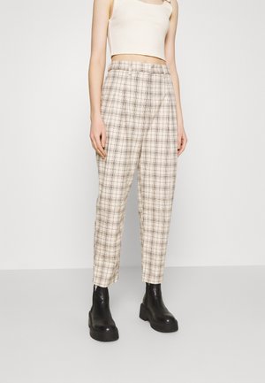 Kvinde iført beige ternede bukser med høj talje, hvid cropped tanktop og sorte chunky ankelstøvler, stående mod en ensfarvet baggrund.