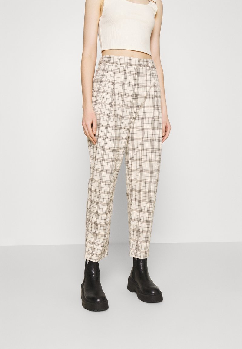 Femme portant un pantalon à carreaux beige taille haute, un débardeur court blanc et des bottines noires épaisses, debout devant un fond uni.
