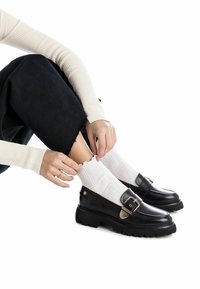 Mocasines de cuero negro con suela gruesa y detalle de hebilla, acompañados de calcetines blancos de canalé, sobre un fondo blanco liso.
