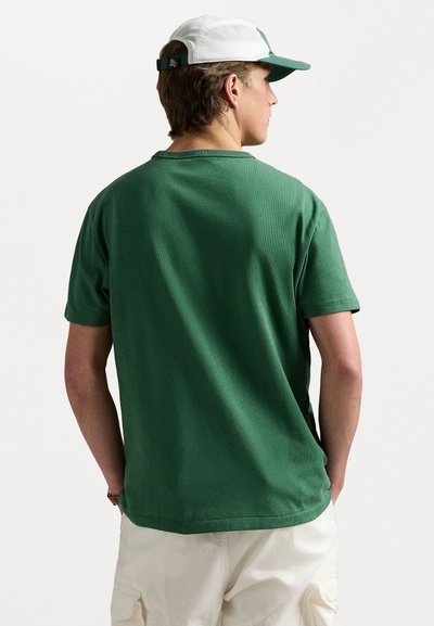Grön t-shirt med kort ärm och strukturerad yta, rund halsringning och avslappnad passform. Bärs med en ljus mössa och shorts.