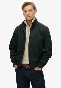 Chaqueta bomber verde oscuro con un cuello alto, cremallera frontal y puños de canalé. Combinada sobre un suéter beige con cremallera y jeans de mezclilla oscuros.