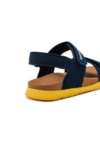 Sandalia de tela azul con suela de goma amarilla, correa ajustable y plantilla texturizada. Presenta un respaldo acolchado y diseño de punta abierta.