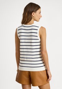 Fehér ujjatlan kötött felső tengerészkék vízszintes csíkokkal, bordázott nyakkivágással és szegéllyel. Barna bőr rövidnadrággal viselve. Egyszerű háttér.