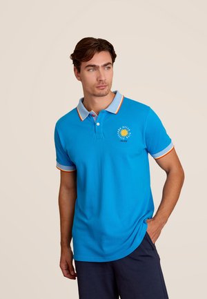 Chemise polo turquoise avec un col rayé comportant des accents rouges, orange et jaunes. Comprend un motif de soleil brodé sur la poitrine.