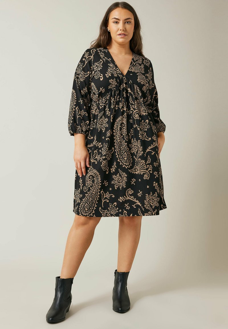 Schwarzes Kleid mit beigem Paisleymuster, V-Ausschnitt und 3/4-Ärmeln. Weicher Stoff mit fließender Silhouette. Getragen mit Ankle Boots.