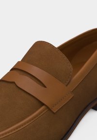 Καφέ μοκασίνι από σουέντ με λείο δερμάτινο λουράκι στην κορυφή και ορατή ραφή κατά μήκος των ακρών.