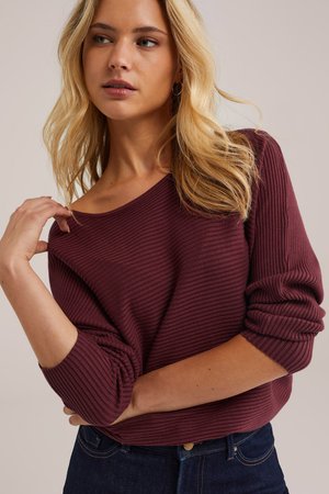 Burgundska rebrasta pulover s širokim izrezom, ohlapnim krojem in zavitimi rokavi, v kombinaciji s temno denimsom. Gladka tekstura, minimalne detajle.