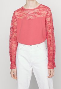 Koraal kleurige top met lange kant sleeves, voorzien van een vaste ronde halslijn en een relaxte pasvorm. Het kant heeft een bloemenpatroon en gerafelde manchetten. Gecombineerd met witte jeans.