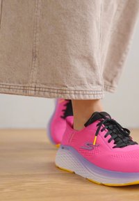 Pink athletic shoes with a textured upper, black laces, and a thick white sole accented by purple and yellow details, paired with a beige skirt.