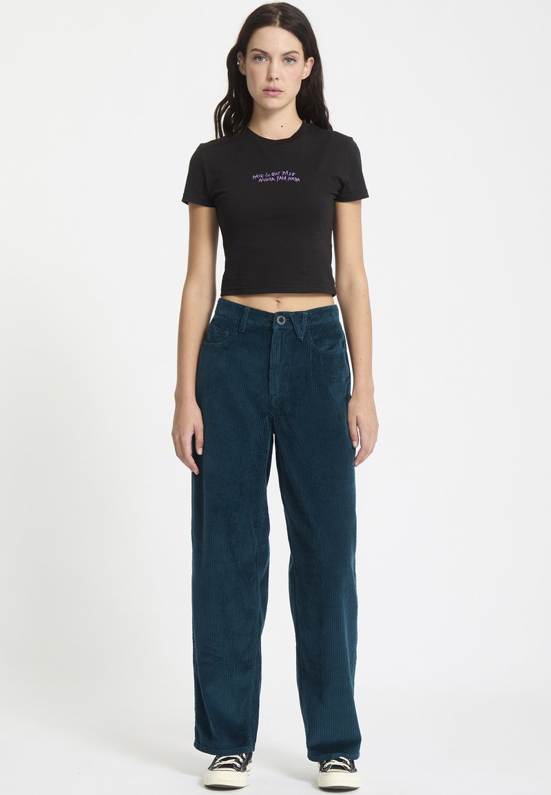 T-shirt noir court avec texte violet, associé à un pantalon large en velours côtelé teal avec fermeture à bouton et poches classiques.