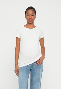 Camiseta de maternidad blanca de canalé con mangas cortas y un escote redondo, que presenta un corte holgado y bordes de las mangas con volantes, combinada con jeans azules.