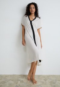 White knee-length dress with short sleeves, black front panel, and gold accents. Soft fabric with a slight drape, featuring a side slit.