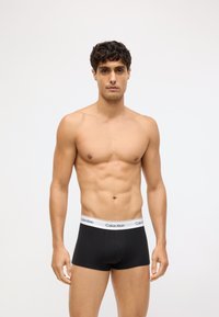 Czarne bokserki z białą gumką z logo Calvin Klein, wykonane z miękkiego materiału, dopasowany krój. Mężczyzna ma muskularną budowę i krótkie kręcone włosy.