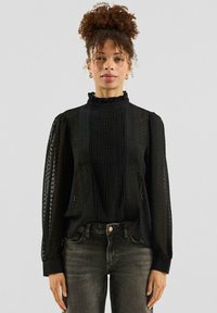 Zwarte blouse met gestructureerde stof, ruches aan de halslijn en geplooide voorkant. Lange mouwen met geplooide manchetten, gecombineerd met donkere jeans.