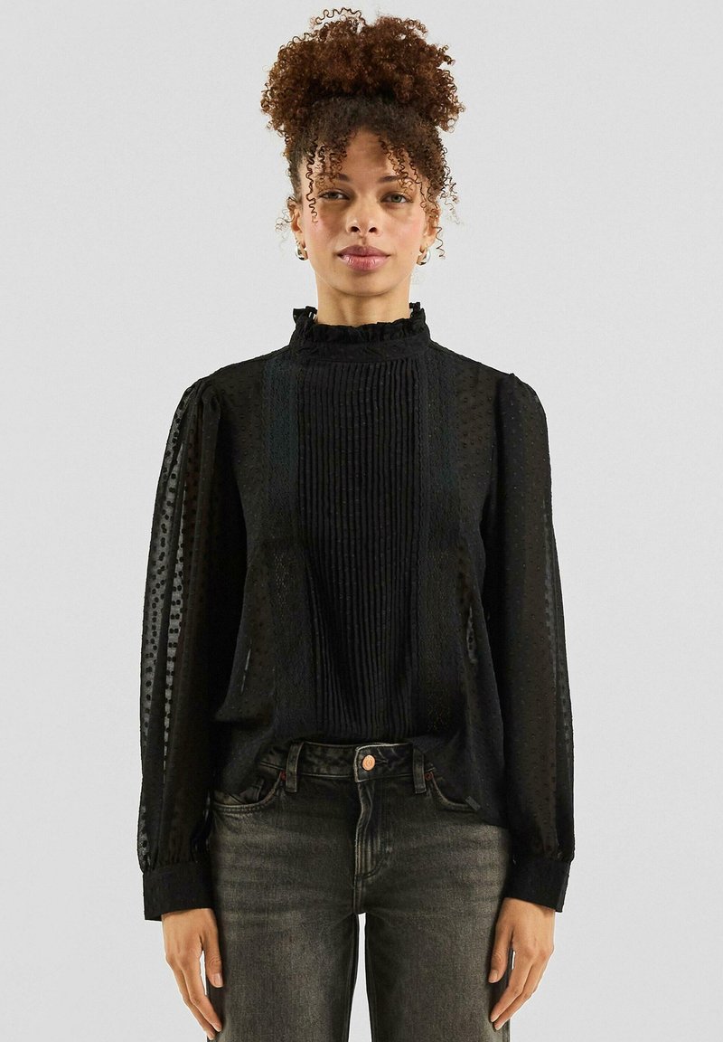 Zwarte blouse met gestructureerde stof, ruches aan de halslijn en geplooide voorkant. Lange mouwen met geplooide manchetten, gecombineerd met donkere jeans.