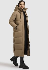 Lungo cappotto imbottito color marrone chiaro con cappuccio, caratterizzato da un design trapuntato, tasche laterali e una zip frontale. Abbinato a stivaletti neri con lacci.