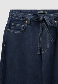 Pantaloni in denim blu scuro con una cintura legata in vita, dotati di tasche frontali con accessori in rame e cuciture gialle a contrasto.