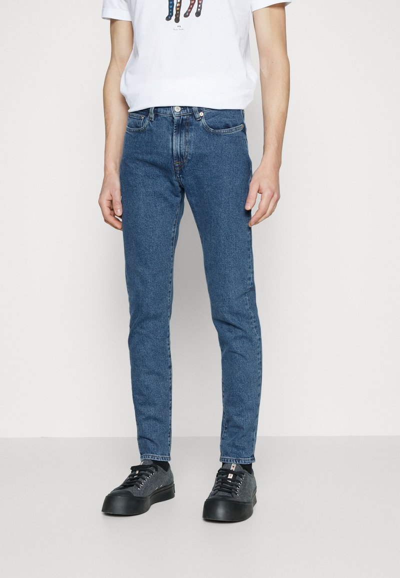 Sittande blå denimjeans med kontrastsömnad, fem-fickorsdesign och en klassisk midja; i kombination med svarta skor och en vit T-shirt.