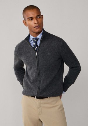 Suéter de cremallera gris oscuro hecho de material suave, con cuello alto y puños acanalados, superpuesto a una camisa a rayas azul y blanca.