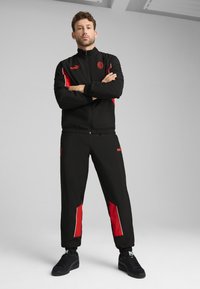 Ensemble de sport noir avec accents rouges, composé d'une veste zippée et d'un pantalon à taille élastique. Fabriqué en tissu lisse et léger.